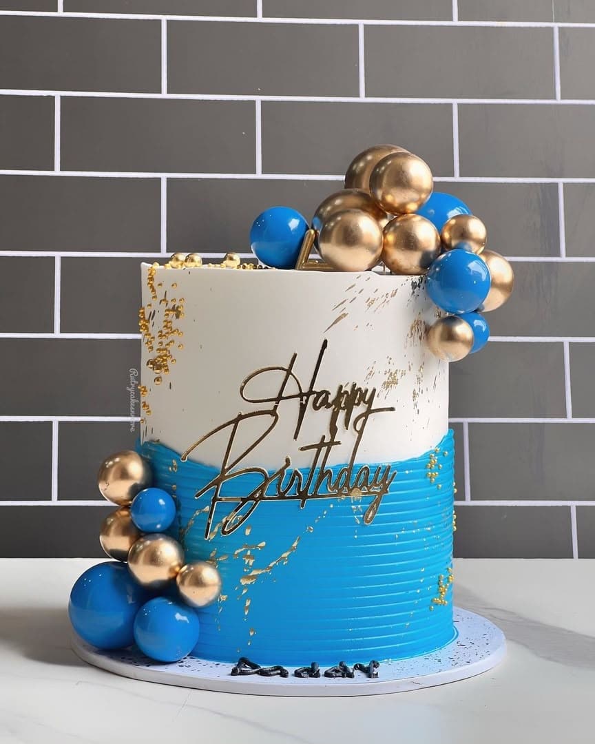 A blue and white birthday cake with gold drip and sphere decorations