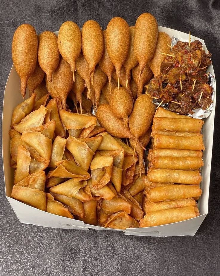 A box of corn dogs, samosas, spring rolls and suya skewers